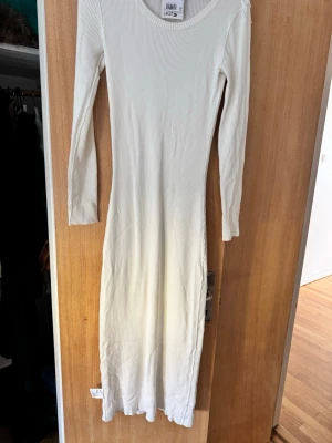 Vit ribbad långärmad klänning - Snygg vit ribbad klänning med lång ärm och rund halsringning. Klänningen är lång och har en smal, kroppsnära passform. Perfekt för dig som gillar en enkel men trendig stil.