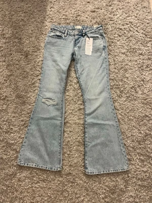 Ljusblå bootcut jeans med slitning - Säljer ett par ljusblå bootcut jeans med slitning på ena låret. Jeansen har klassisk femficksmodell, låg midja och vida ben. Materialet är denim i bomull och de har en snygg vintagekänsla. Perfekta för dig som gillar retrostil och vill ha ett par trendiga jeans. Strl 36 men passar även 34! helt nya med prislapp💕