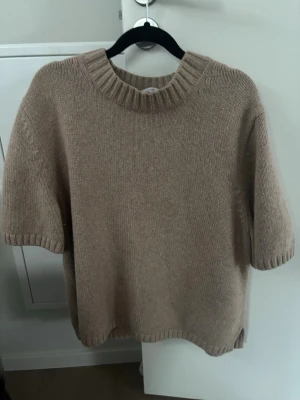 Beige kashmir tröja med kort ärm - En enkel och stilren beige stickad tröja med korta ärmar och bred ribbad halsringning i kashmir. Tröjan har en rak passform och ribbade muddar vid ärmslut och nederkant. Perfekt för lager på lager och en avslappnad look.