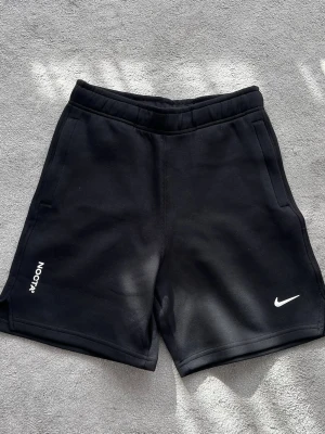 Svarta NOCTA x Nike shorts - Snygga svarta shorts från NOCTA x Nike med vit logga på ena benet och NOCTA-tryck på det andra. De har en relaxed passform, elastisk midja och sidofickor. Perfekta för sommaren och träning, tillverkade i mjuk bomullsmix.