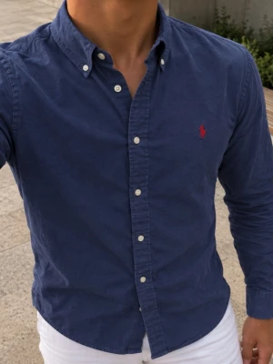 Marinblå skjorta från Ralph Lauren - Snygg marinblå skjorta från Ralph Lauren med klassisk button-down krage och vita knappar. Skjortan har lång ärm och den ikoniska röda logotypen broderad på bröstet. Tillverkad i mjuk bomull för en bekväm känsla.