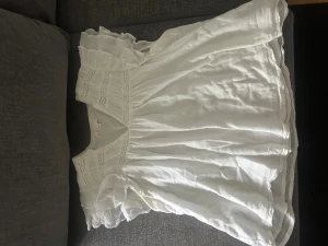 Vit blus med volang från Gina Tricot - Söt vit blus från Gina Tricot med volangdetaljer på ärmarna och v-ringad hals. Tillverkad i ett lätt och luftigt material, perfekt för varma dagar. Blusen har fina broderade detaljer upptill och en lös passform.
