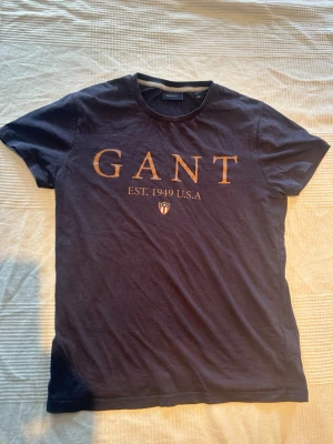 Marinblå GANT t-shirt med tryck - Snygg marinblå t-shirt från GANT med stort tryck framtill i guldtext och logga. Klassisk rund halsringning och korta ärmar. Tillverkad i mjuk bomull som känns skön mot huden. Perfekt för en clean och stilren look.