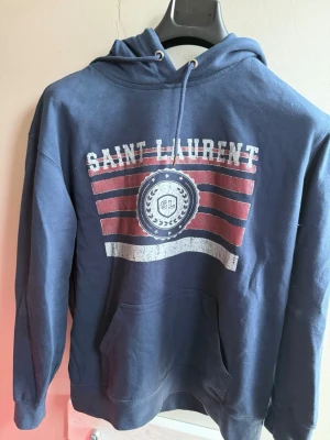 Blå Saint Laurent hoodie med tryck - Mörkblå hoodie från Saint Laurent med huva och dragsko. Stor ficka framtill och ett stort tryck på bröstet med texten 'Saint Laurent' och röda, vita detaljer. Tillverkad i mjukt bomullsmaterial, perfekt för en avslappnad stil.