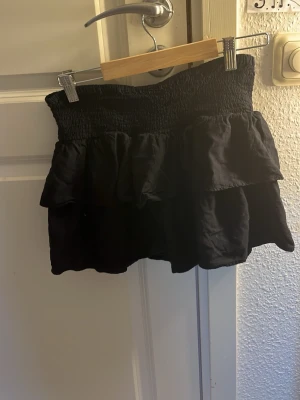 Mörkblå  volangkjol med smock - Supersöt mörkblå kortkjol med bred smock i midjan och dubbla volanger. Kjolen har en luftig och flowy känsla, perfekt för att styla med crop tops eller stickade tröjor. Passar dig som gillar en feminin och trendig look.