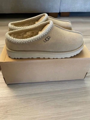  UGG Tasman Mustard seed  - Mysiga beige tofflor från UGG i mocka med fluffigt fårskinnsfoder. Klassisk slip-on modell med grov sula och dekorativt mönster runt öppningen. Perfekta för att hålla fötterna varma hemma.