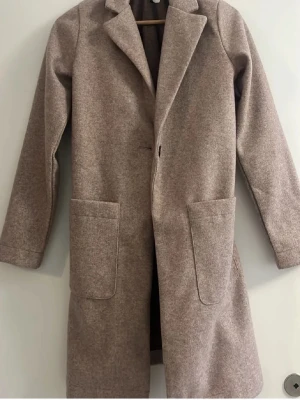 Beige ullkappa med stora fickor - Stilren beige kappa i mjukt, lent material med rak passform och klassisk krage. Kappan har två stora utanpåfickor och enkel knäppning framtill. Ofodrad, passar för höst eller vår. Använd typ två gånger i nån timme var. Öppen för prisförslag