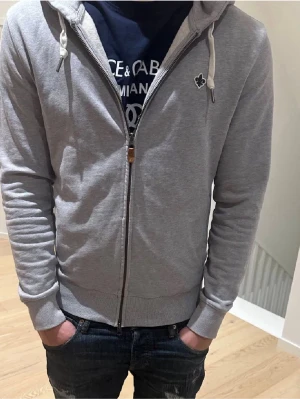 Ljusgrå zip hoodie från Morris - Säljer en ljusgrå hoodie från Morris med dragkedja framtill och vit dragsko i huvan. Hoodien har en liten broderad logga på bröstet och ribbade muddar vid ärmar och nederkant. Tillverkad i mjukt bomullsmaterial för skön känsla.