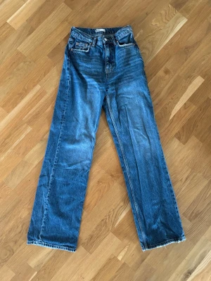 Blå raka jeans med hög midja - Säljer ett par klassiska blå jeans med rak passform och hög midja. Jeansen har fem fickor, bälteshällor och råa kanter vid bensluten för en trendig look. Tillverkade i kraftigt denimtyg som ger en snygg vintagekänsla.
