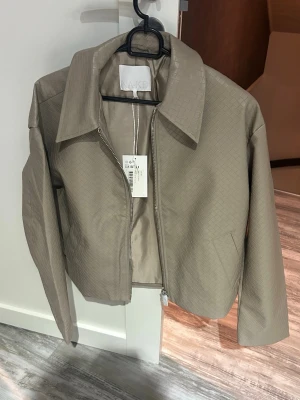 Beige skinnjacka med struktur från NA-KD - Snygg beige översized skinnjacka från NA-KD med diskret rutmönstrad struktur. Jackan har rak passform, bred krage och dragkedja framtill. Perfekt för dig som vill ha en stilren och trendig look. Storlek är xxs men passar som xs/s.