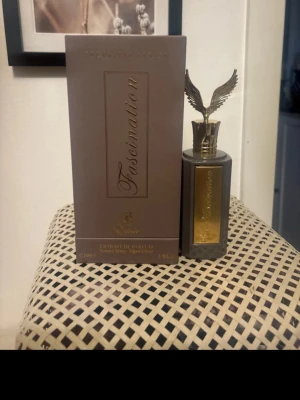 Fascination Extrait de Parfum - Lyxig parfym i en elegant grå flaska med gulddetaljer och en unik kork formad som vingar. Flaskan har en exklusiv känsla och kommer i matchande ask med guldfärgad text. Perfekt för dig som vill sticka ut med din doft.