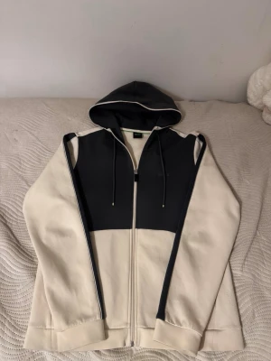 Svart-beige hoodie från BOSS - Snygg hoodie från BOSS i svart och beige med dragkedja framtill och justerbar huva. Hoodien har en diskret BOSS-logga på bröstet. Tillverkad i mjukt material som känns skönt mot huden. Hoodien är använd fåtal gånger, pris kan diskuteras.