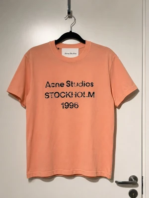 Persikofärgad Acne Studios t-shirt - Säljer en persikofärgad t-shirt från Acne Studios med svart tryck på bröstet där det står 'Acne Studios STOCKHOLM 1996'. T-shirten har klassisk passform och korta ärmar. Perfekt för dig som gillar stilrena plagg med en twist.