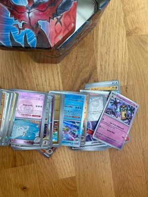 Samling Pokémonkort med plåtlåda - Stor samling Pokémonkort i olika färger och motiv, inklusive flera holografiska och vanliga kort. Korten kommer i en färgglad plåtlåda med Pokémon-tryck. Perfekt för dig som vill utöka din samling eller börja spela Pokémon TCG. Endast holos och revers holos kan även finnas äldre kort 