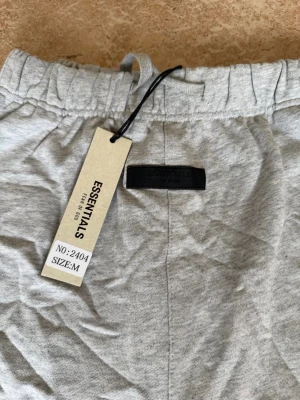 Grå shorts från Essentials Fear of God - Snygga grå shorts från Essentials Fear of God med elastisk midja och logga tryckt på vänster ben. Tillverkade i mjuk bomull för maximal komfort. Perfekta för en avslappnad och trendig stil.