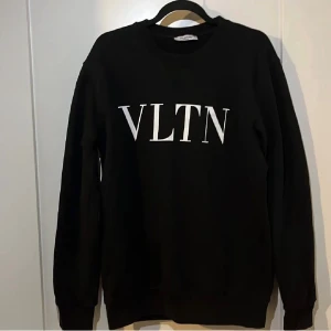 Svart sweatshirt från Valentino - Säljer en svart sweatshirt från Valentino med VLTN-tryck i vitt på bröstet. Tröjan har rund halsringning, långa ärmar och ribbade muddar vid ärmslut och nederkant. Perfekt för en stilren och trendig look.