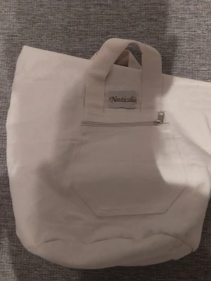 Vit shoppingväska från Nauticalia - Stilren vit shoppingväska i canvas från Nauticalia. Väskan har två korta handtag, en praktisk ytterficka med dragkedja på framsidan och en öppen ficka på baksidan. Perfekt för att bära med sig allt man behöver under dagen.