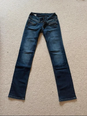 Mörkblå raka jeans med fickdetaljer - Snygga mörkblå jeans med raka ben och klassiska fickor fram och bak. Jeansen har diskreta slitningar och coola sömdetaljer på bakfickorna. Perfekta för en avslappnad och trendig look. Materialet är denim i bomull.