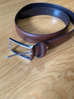 Brunt läderbälte med silverfärgat spänne - Snyggt brunt bälte i äkta läder med klassiskt silverfärgat metallspänne. Bältet har en stilren design och passar perfekt till jeans eller kostymbyxor. Enkel att justera i längden tack vare flera hål.