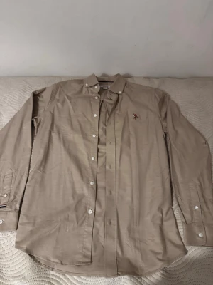 Beige skjorta med broderad logga - Snygg beige skjorta i bomull med klassisk krage och knappar framtill. Skjortan har lång ärm och en broderad logga på bröstet i färgerna röd, vit och brun. Perfekt för en clean och stilren look. Ny utan prislapp.