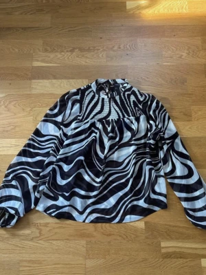 Zebramönstrad blus från Lindex - Snygg zebramönstrad blus från Lindex i svart och vitt. Blusen har hög krage, smock upptill och långa ärmar med resår vid ärmslut. Tillverkad i ett lätt och luftigt material som ger en cool look.