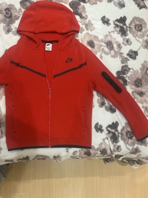  Nike Tech Fleece - Passar S.Snygg ÄKTA röd Nike Tech Fleece hoodie med dragkedja framtill och svarta detaljer. Hoodien har en huva, två sidofickor och en unik ficka på ärmen. Tillverkad i ett mjukt och stretchigt material som ger en sportig vibe. Nike-logga på bröstet och cool design med svarta linjer. ANVÄND BARA 2-3 GÅNGER, JÄTTEFRÄSCH.