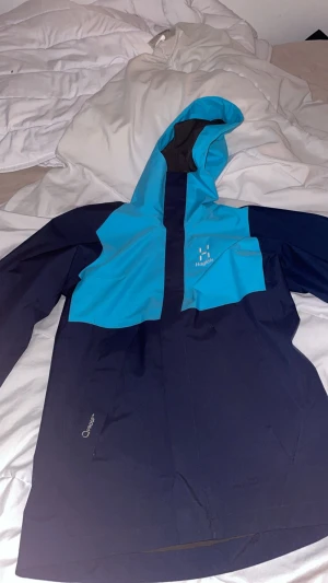 Blå och marinblå vindjacka Haglöfs XS - Snygg vindjacka från Haglöfs i färgerna blå och marinblå. Jackan har huva, dragkedja framtill och meshfoder på insidan. Perfekt för blåsiga dagar och outdoor-äventyr. Modellen är för kvinnor och har en sportig look.