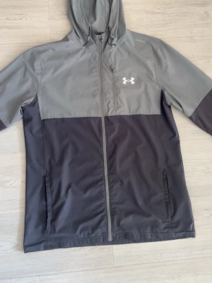 Grå vindjacka från Under Armour - Snygg tvåfärgad vindjacka från Under Armour i ljusgrått och mörkgrått. Jackan har huva, hel dragkedja, två sidofickor och en bröstficka med dragkedja. Perfekt för blåsiga dagar och sportiga outfits. Under Armour-logga på bröstet.