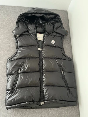 Svart dunväst från Moncler med huva - Snygg svart dunväst från Moncler med quiltad design och avtagbar huva. Västen har två dragkedjeförsedda fickor och Moncler-logga på bröstet. Perfekt för lager-på-lager och håller dig varm under kyliga dagar. Aldrig använd enbart testad