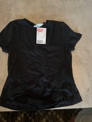 Svart t-shirt med snörningsdetalj - En svart t-shirt med korta ärmar och snygga snörningsdetaljer på sidorna nedtill. T-shirten har en klassisk rund halsringning och är tillverkad i ett mjukt material som känns skönt mot huden. Perfekt för en avslappnad och stilren look.