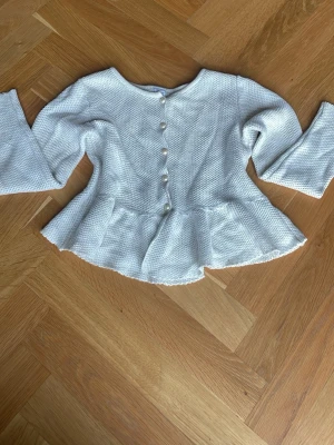 Vit peplumkofta med pärlknappar - Supersöt vit kofta med peplumskärning och pärlknappar. Materialet är stickat med diskret glittrig tråd som ger en extra touch. Volang längst ner