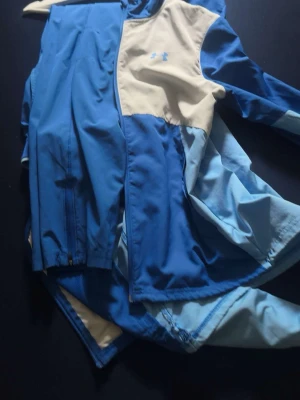 Blå och vit tracksuit  från Under Armour - Snygg tracksuit  från Under Armour i olika blå nyanser och beige panel upptill.  framtill och sportig design med logga på bröstet. Tillverkad i lätt och funktionellt material som passar perfekt för aktiva dagar.passar även vissa s den är äkta tar byten beror på vad och pris kan diskuteras 