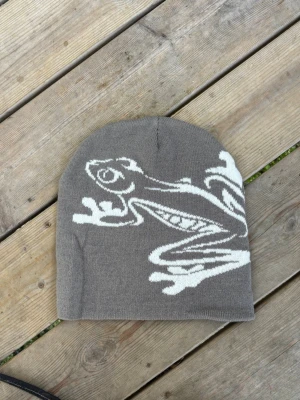 LjusGrå mössa med vit groda Beanie  - Säljer en grå stickad mössa med en stor vit groda som motiv på sidan. Mössan har en klassisk form och är tillverkad i mjukt material som passar perfekt för kyliga dagar. Snygg och unik accessoar för dig som vill sticka ut lite extra.
