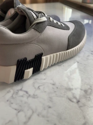 Grå och vita sneakers med sicksack-sula - Snygga sneakers i grått och vitt med ovandel i textil och mockaliknande material. Skorna har vita snören och en unik sicksack-mönstrad sula med svart detalj på sidan. Perfekt för dig som gillar modern och stilren design.