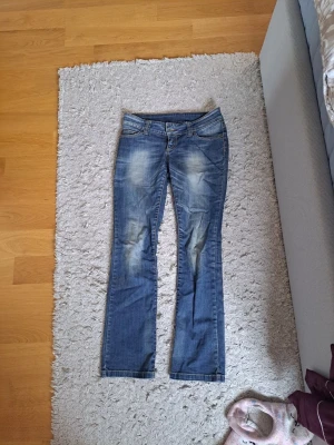Blå bootcut jeans med slitningar - Säljer ett par blå bootcut jeans med snygga slitningar och tvättade detaljer framtill. Jeansen har låg midja, klassisk femficksmodell och stängs med knapp och dragkedja. Perfekta för en avslappnad och trendig look.