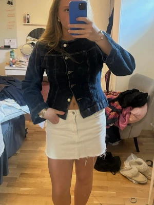 Mörkblå figursydd jeansjacka - Säljer en mörkblå jeansjacka med figursydd passform och dekorativa sömmar. Jackan har rundad halsringning, långa ärmar och knäpps med fem metallknappar framtill. Perfekt för dig som vill ha en unik twist på den klassiska jeansjackan.