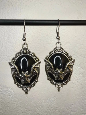 Gotiska fladdermusörhängen i viktoriansk stil med silver- och svartfärgad cameo – vampyr, witchy, alternativ stil - Stora örhängen i gotisk/viktoriansk stil med svarta cameos och fladdermöss i silverfärgad metall. Ett riktigt statement-smycke för dig som gillar goth, alternative, witchy, dark academia eller vampyrstil. Toppskick. Allergivänliga krokar och nya silikonstoppar medföljer. Mått ca 6 × 2,5 cm. Bundla gärna – du får rabatt vid köp av flera annonser.