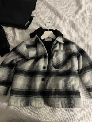 Rutig overshirt med huva - Snygg overshirt i svartvitt rutigt mönster med svart huva. Jackan har knäppning framtill, en bröstficka och är tillverkad i ett mjukt tyg som ger en avslappnad look. Perfekt att slänga över en hoodie för extra stilpoäng.