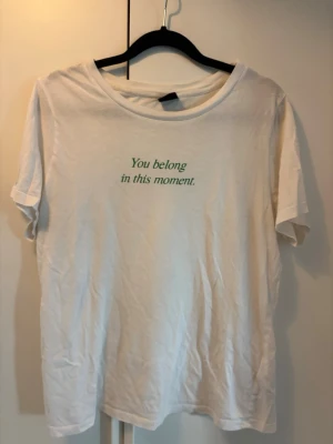 Vit t-shirt med texttryck - Vit t-shirt i bomull med rund hals och korta ärmar. På bröstet står det 'You belong in this moment.' i grön text. Enkel och clean design som passar till det mesta.