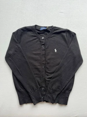 Ralph Lauren Tröja - Klassisk svart tröja med rund hals och knäppning framtill från Polo Ralph Lauren. Tröjan har den ikoniska vita logobrodyr på bröstet och ribbade muddar vid ärmslut och nederkant. Perfekt att slänga över en t-shirt för en clean look.