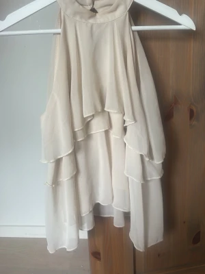 Beige volang topp med omlottkänsla - Säljer en superfin beige topp med flera lager volanger och omlottkänsla. Toppen är ärmlös och har en luftig, flowy passform som ger en romantisk vibe. Perfekt att styla med jeans eller kjol för en trendig look.
