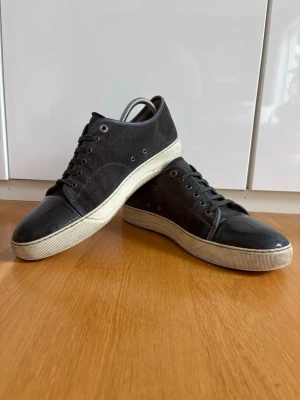 Lanvin skor - Snygga svarta sneakers i mocka med cool lackad tå och vit sula från Lanvin. Skorna har klassisk låg profil, svarta skosnören och metallöljetter. Perfekta för dig som gillar en stilren men ändå unik look. Storlek 44