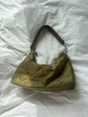 Olivgrön quiltad axelväska - Snygg olivgrön handväska i quiltad design från ”Unlimit Bag” med kort axelrem och dragkedja. Väskan är gjord i glansigt tyg med guldfärgat innerfoder och har en liten innerficka med dragkedja. Perfekt för dig som vill ha en trendig accessoar till din outfit.