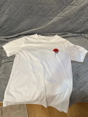 Vit t-shirt med röd rosdetalj - En helt ny tshirt i Storlek M. Säljer en stilren vit t-shirt med en liten broderad röd ros på bröstet. T-shirten har rund halsringning. Perfekt för dig som gillar enkla plagg med en twist.