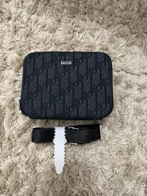 Dior svart monogram plånbok - Snygg svart plånbok från Dior med klassiskt monogrammönster och silverfärgad dragkedja. Plånboken har flera kortfack och ett större fack inuti samt en avtagbar handledsrem. Perfekt för dig som vill ha både stil och funktion.