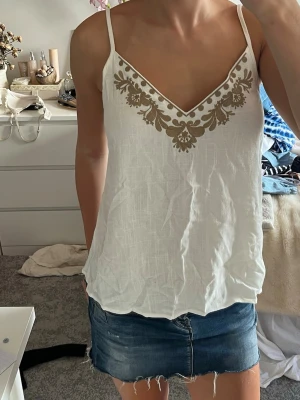 Vit linnetopp med broderi - Säljer en vit linnetopp med tunna axelband och v-ringning. Toppen har ett snyggt beige broderat mönster vid urringningen som ger en bohemisk vibe. Perfekt till sommaren och lätt att matcha med jeans eller kjol.