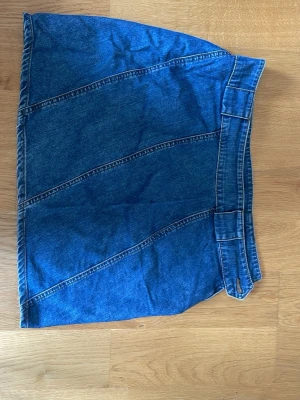 Blå jeanskjol med bred linning - Säljer en blå jeanskjol i kort modell med bred linning och markerade sömmar i diagonala paneler. Kjolen har en klassisk denimlook och är tillverkad i bomull. Perfekt för dig som gillar en enkel men snygg stil.