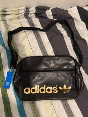 Svart axelväska från Adidas - Snygg svart axelväska från Adidas med justerbar rem och stort guldfärgat Adidas-tryck på framsidan. Väskan har en dragkedjeficka framtill och är gjord i ett glansigt konstlädermaterial. Perfekt för att bära med sig det viktigaste och ger en sportig retrostil.