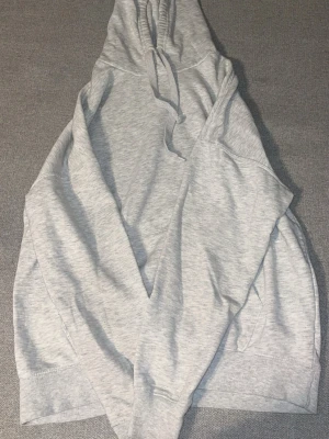 Ljusgrå basic hoodie med huva - En stilren ljusgrå hoodie med huva och dragsko. Hoodien har ribbade muddar vid ärmslut och nederkant samt en klassisk känguruficka framtill. Perfekt för en avslappnad och enkel look. Materialet känns mjukt och bekvämt.