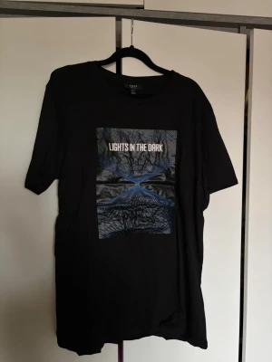 Svart t-shirt från SMOG med tryck - Cool svart t-shirt från SMOG med trycket 'LIGHTS IN THE DARK' och grafiskt motiv i blått och vitt på bröstet. 